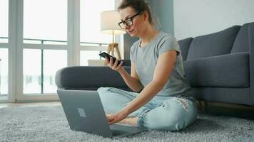 casualmente vestido mulher sentado em tapete com computador portátil e Smartphone e trabalhando dentro acolhedor sala. controlo remoto trabalhos lado de fora a escritório video