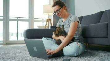 mine de rien habillé femme avec des lunettes est assis sur une tapis avec une ordinateur portable, détient sur sa les genoux et coups une duveteux chat et travaux dans une confortable chambre. éloigné travail à l'extérieur le Bureau video