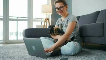 casualmente vestido mulher com óculos senta em uma tapete com uma computador portátil, detém em dela joelhos e golpes uma fofo gato e trabalho dentro uma acolhedor sala. controlo remoto trabalhos lado de fora a escritório video