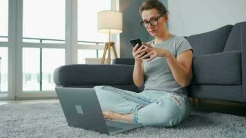 mine de rien habillé femme séance sur tapis avec portable et téléphone intelligent et travail dans confortable chambre. éloigné travail à l'extérieur le Bureau video