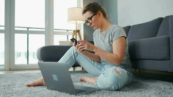 mine de rien habillé femme séance sur tapis avec portable et téléphone intelligent et travail dans confortable chambre. éloigné travail à l'extérieur le Bureau video