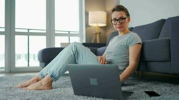 Casually dressed woman with glasses is sitting on carpet with laptop in cozy room. She communicates and works remotely. Remote work outside the office video