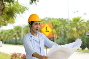 Asian man malay worker engineer management hard hat safety helmet at construction site reading seeing plan blueprint look surprised photo