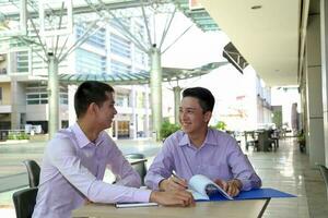 Young south east Asian middle eastern man business colleague outdoor cafe chair table meeting talking discussing file paper photo