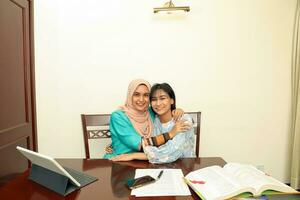 Two young Asian Malay Muslim woman wearing headscarf at home office student sitting at table phone computer book document selfie self portrait hug care affection photo