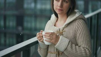 Kaukasisch vrouw blijft Aan balkon gedurende sneeuwval met kop van heet koffie of thee. ze looks Bij de sneeuwvlokken en ademt video