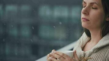 Caucasian woman stays on balcony during snowfall with cup of hot coffee or tea. She looks at the snowflakes and breathes video