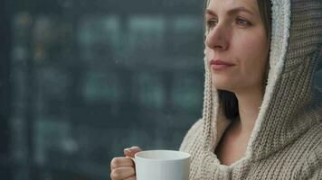 Kaukasisch vrouw blijft Aan balkon gedurende sneeuwval met kop van heet koffie of thee. ze looks Bij de sneeuwvlokken en ademt video