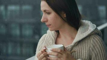 caucasiano mulher permanece em sacada durante queda de neve com copo do quente café ou chá. ela parece às a flocos de neve e respira video