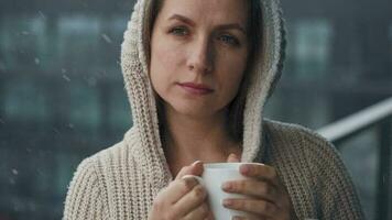 Kaukasisch vrouw blijft Aan balkon gedurende sneeuwval met kop van heet koffie of thee. ze looks Bij de sneeuwvlokken en ademt video