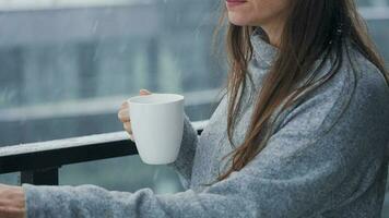 Kaukasisch vrouw blijft Aan balkon gedurende sneeuwval met kop van heet koffie of thee. ze looks Bij de sneeuwvlokken en ademt video