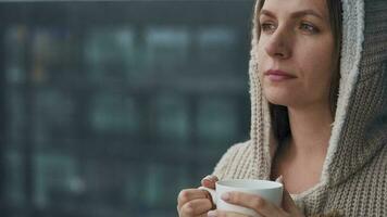 Kaukasisch vrouw blijft Aan balkon gedurende sneeuwval met kop van heet koffie of thee. ze looks Bij de sneeuwvlokken en ademt video