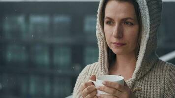 Kaukasisch vrouw blijft Aan balkon gedurende sneeuwval met kop van heet koffie of thee. ze looks Bij de sneeuwvlokken en ademt video