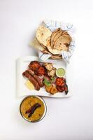 Mixed grill kebab chicken mutton beef prawn fish mint sauce lemon pickled onion bread basket chapati naan tandoori paratha dal lentil fry tadka copper pot on white dish over white background photo