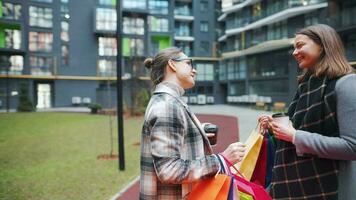 Two happy women stand with shopping bags and takeaway coffee after a successful shopping and talk with interest among themselves. Modern buildings on background. Slow motion video