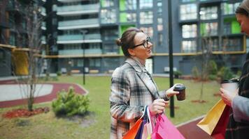 Two happy women stand with shopping bags and takeaway coffee after a successful shopping and talk with interest among themselves. Modern buildings on background. Slow motion video