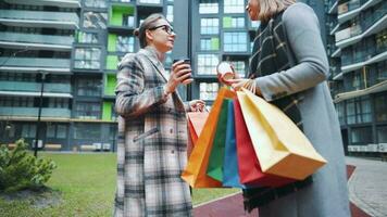 zwei glücklich Frauen Stand mit Einkaufen Taschen und wegbringen Kaffee nach ein erfolgreich Einkaufen und sich unterhalten mit Interesse unter sich. modern Gebäude auf Hintergrund. schleppend Bewegung video