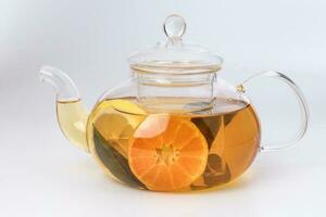 Liquid tea lemon orange slice green leaf cinnamon stick in transparent glass teapot kettle on white background photo