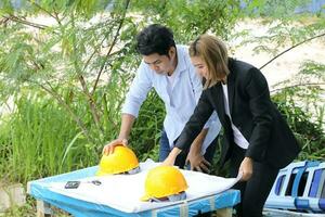 Man woman Malay Chinese worker engineer management hard safety hat helmet at construction site reading seeing plan on outdoor makeshift table photo