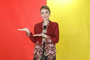 Asian woman traditional maroon kebaya sharong on red yellow paper background posing show display palm of hand photo