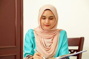 young Asian Malay Muslim woman wearing headscarf at home office student sitting at table read write book document note study smile happy look photo