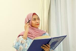 young Asian Malay Muslim woman wearing headscarf at home office next to window look at book document study think write photo