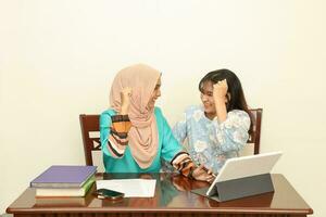 Two young Asian Malay Muslim woman wearing headscarf at home office student sitting at table talk mingle computer book document study discuss fist in air happy victory photo