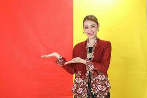 Asian woman traditional maroon kebaya sharong on red yellow paper background posing show display palm of hand photo