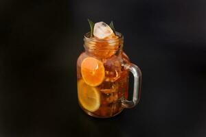 Liquid ice lemon orange tea with slice green leaf cinnamon stick in transparent glass jar mug on black background photo
