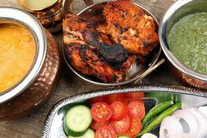 Spicy red Chicken grilled tikka tandoori nan bread green spinach curry yogurt sauce dal tomato cucumber onion salad set in metal stainless steel brass copper pot on rustic vintage wood background photo