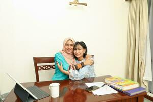Two young Asian Malay Muslim woman wearing headscarf at home office student sitting at table phone computer book document selfie self portrait hug care affection photo