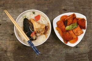 Fish head noodles vermicelli milk soup in white bowl pan friend salmon fish fillet on triangular plate chopstick spoon on rustic wooden background photo