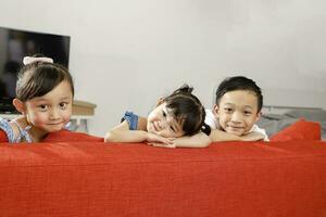 Tres pequeño niño chico niña hermano hermana contento sonrisa mirando terminado el sofá foto