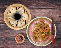 Home style dim sum dumpling in bamboo steamer chili flake oil sauce Stair fried noodle with prawn seafood tofu colorful pepper on oriental style plate chopsticks over rustic wooden background photo
