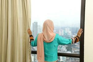 joven asiático malayo musulmán mujer vistiendo Pañuelo a hogar oficina estudiante a ventana Mira fuera de a el ciudad edificio foto