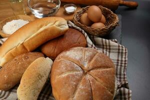 Freshly baked bread loaf bun roll round long mix verity wrapped in checkered kitchen fabric napkin towel wheat flower oil water salt eggs rolling pin slate stone over black background photo