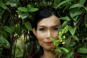 One Asian Malay woman posing look forward towards camera through green leaf outdoor green park photo