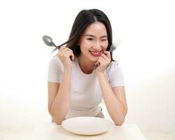 Beautiful young south east Asian woman pretend acting posing holding empty fork spoon in hand eat taste look see white plate on table white background smile happy exited wink photo