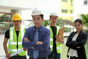 South East Asian young Malay Chinese man woman safety helmet vest formalwear stand at construction site blueprint plan clipboard look at camera photo