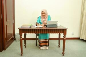 joven asiático malayo musulmán mujer vistiendo Pañuelo a hogar oficina estudiante sentar a mesa computadora libro archivo papel estudiar trabajo foto