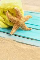 Orange starfish with yellow towel on blue wooden board of pier or sun lounger on sand of beach. Vacation, travel, weekend. Copy space photo