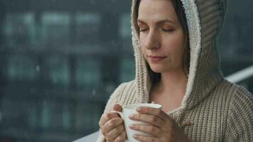 Kaukasisch vrouw blijft Aan balkon gedurende sneeuwval met kop van heet koffie of thee. ze looks Bij de sneeuwvlokken en ademt video