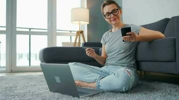 mujer con lentes es sentado en el alfombra y hace un en línea compra utilizando un crédito tarjeta y teléfono inteligente en línea compras, estilo de vida tecnología video