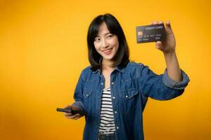 Portrait beautiful young asian woman happy smile dressed in denim jacket showing smartphone and credit card isolate on yellow studio background. Shopping online smartphone application concept photo