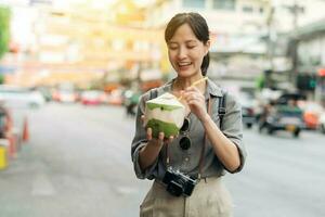 Happy young Asian woman backpack traveler drinking a coconut juice at China town street food market in Bangkok, Thailand. Traveler checking out side streets. photo