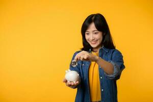 Happy young asian woman wearing yellow t-shirt denim shirt saving digital crypto currency to piggy bank isolated on yellow background. Digital money saving investment concept. photo