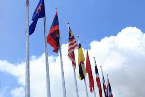 muchas de las banderas de la asean en los coloridos colores soplados por la fuerza del viento ondeando en un poste frente a un hotel en tailandia sobre un fondo con nubes y cielos azules. foto