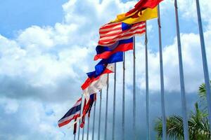 muchas de las banderas de la asean en los coloridos colores soplados por la fuerza del viento ondeando en un poste frente a un hotel en tailandia sobre un fondo con nubes y cielos azules. foto