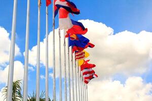 muchas de las banderas de la asean en los coloridos colores soplados por la fuerza del viento ondeando en un poste frente a un hotel en tailandia sobre un fondo con nubes y cielos azules. foto