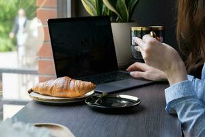 Croissant and female hand with cup of coffee, laptop in cafe. Freelance concept. photo